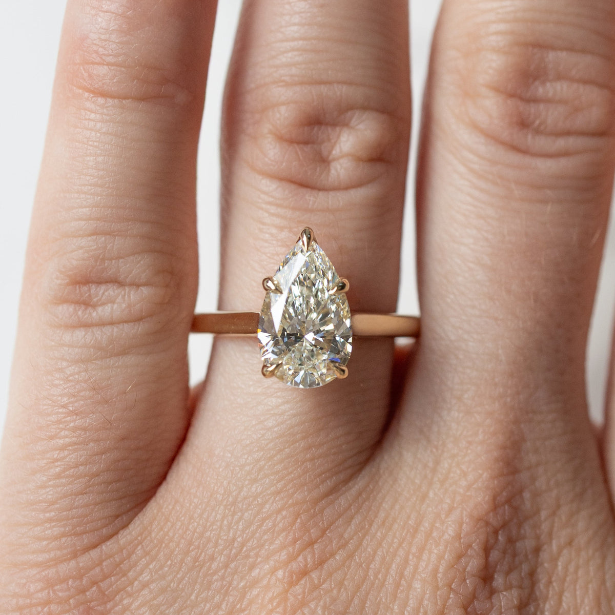 A pear cut diamond engagement ring in a yellow gold setting shown on a left ring finger along a white backdrop.
