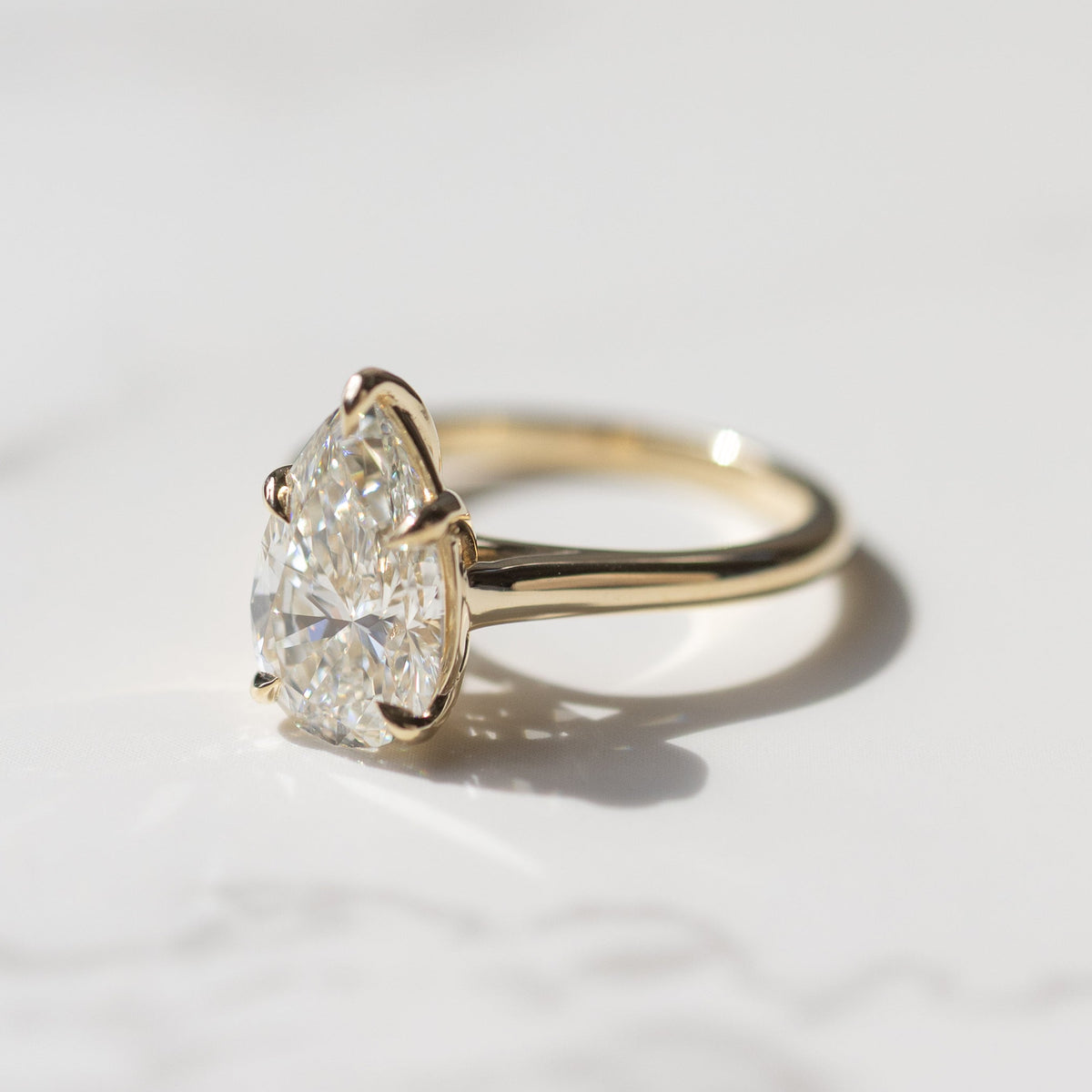 A pear cut diamond engagement ring sitting on a white marble backdrop with soft grey shadows.