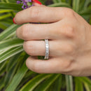 A close up of a hand holding a purple flower with green leaves. On the hand is a white gold diamond wedding band with emerald cut diamonds.