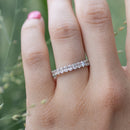 A close-up of a hand wearing a white gold eternity ring set with petite oval diamonds, with a soft-focus green outdoor background.