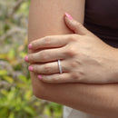 A close-up lifestyle shot of a woman's hand resting on her elbow, wearing a thin white gold band ring set with small oval diamonds. The background is a soft-focus outdoor garden with green foliage and purple flowers.