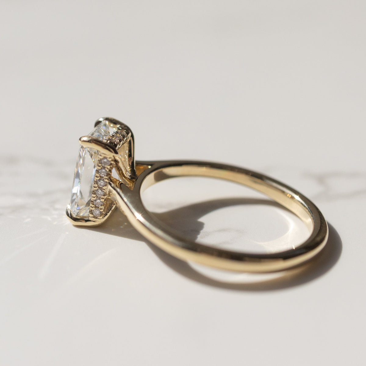 A side profile shot of a large diamond engagement ring with a hidden halo along the gallery rail. The ring is yellow gold and sitting on a moody white backdrop with shadows.