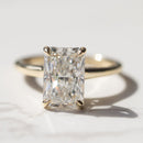 A macro shot of a radiant cut diamond engagement ring. The ring is set in yellow gold and is sitting on a white marble backdrop.