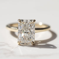 A macro shot of a radiant cut diamond engagement ring. The ring is set in yellow gold and is sitting on a white marble backdrop.