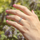 An on-hand shot of a white gold channel set round diamond band. Behind the hand is blurry green and purple flowers.
