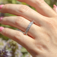 An on-hand shot of a channel set round diamond band set in white gold. Behind the hand are green and purple flowers.