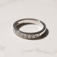 A white gold wedding ring with a row of clear channel-set diamonds, photographed on a light marble surface with a soft shadow.