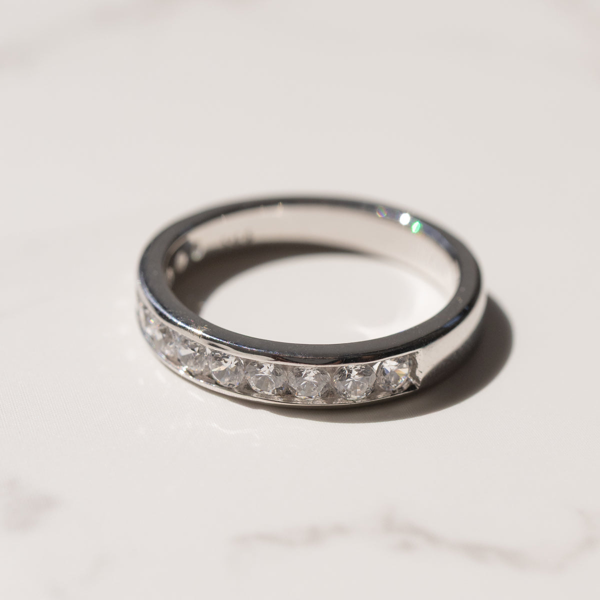 A white gold wedding ring with a row of clear channel-set diamonds, photographed on a light marble surface with a soft shadow.