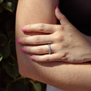An on-hand shot of a white gold round diamond band. The hand is resting on the model's other arm with green leaves in the backdrop.