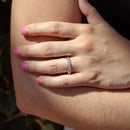 An on-hand model shot of a round diamond eternity band. The hand is resting on the model's other arm and there are dark green leaves in the background.