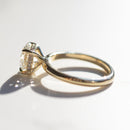 The side profile of an engagement ring that has a round center diamond and hidden halo along its gallery rail. The ring is yellow gold and is sitting on a white marble backdrop.