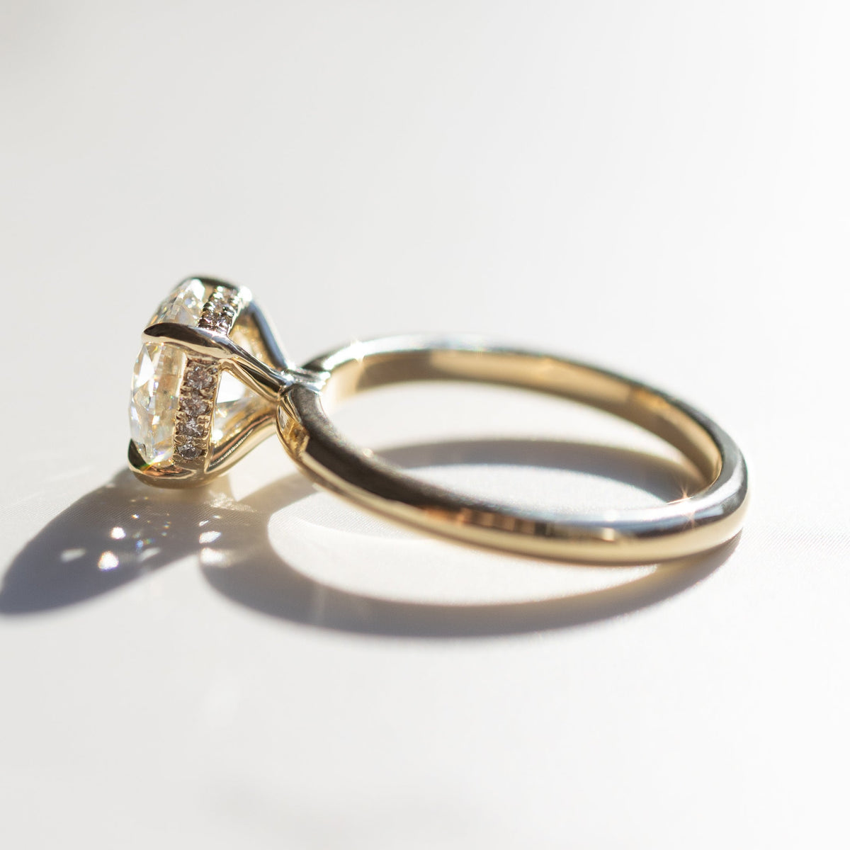 The side profile of an engagement ring that has a round center diamond and hidden halo along its gallery rail. The ring is yellow gold and is sitting on a white marble backdrop.