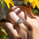 A close up shot of a hand among yellow flowers and green leaves. Highlighted is a large round cut diamond engagement ring sitting on the hand's left ring finger.