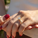 Two hands with red nail polish holding. On one hand is a large round engagement ring with a hidden halo set in yellow gold. Behind the hands are a white top and green foliage.