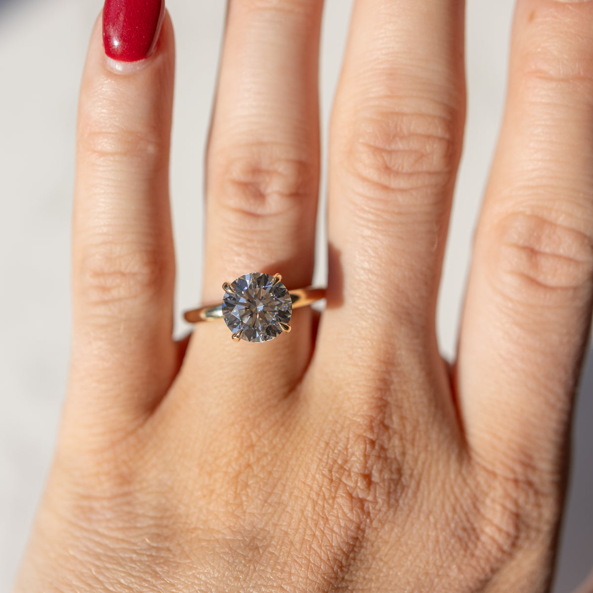 A close up, on-hand shot of a round cut diamond engagement ring. The ring has a large diamond in the center with prongs surrounding it in a yellow gold setting.