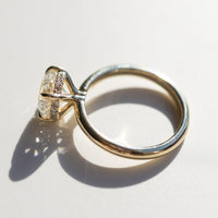 A close up image of a round cut diamond engagement ring on its side. The ring is yellow gold and has a row of small round diamonds set around its gallery rail. The ring is sitting on a white marble backdrop.