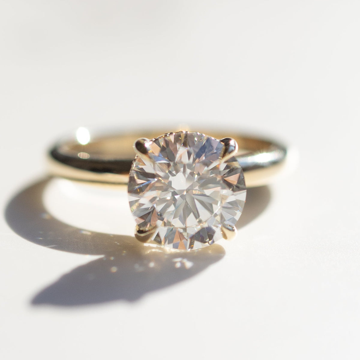 A macro shot of a round diamond engagement ring set in yellow gold. The center diamond is large and prong set. The ring is sitting on a white marble backdrop.