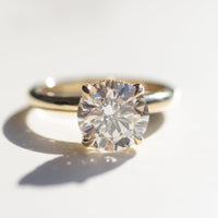 A macro shot of a round diamond engagement ring set in yellow gold. The center diamond is large and prong set. The ring is sitting on a white marble backdrop.