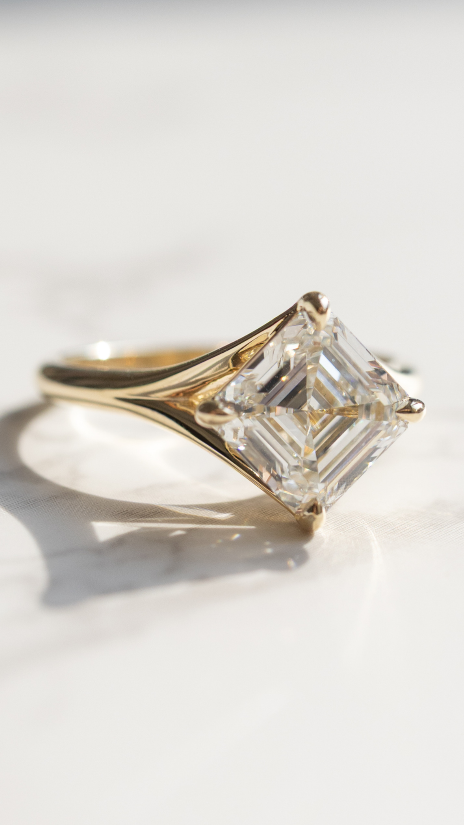 A yellow gold asscher cut diamond ring sitting on a white marble backdrop with shadows.