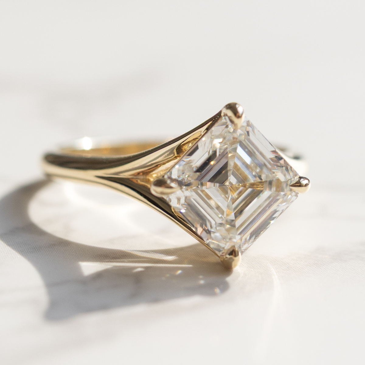 A large asscher cut diamond engagement ring sitting on a white marble backdrop in sunlight. The ring is set in yellow gold.