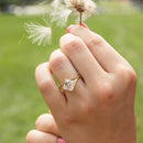 An on-hand shot of a kite set asscher engagement ring against a green field backdrop. The hand is holding a flower blowing in the wind.
