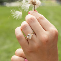 An on-hand shot of a kite set asscher engagement ring against a green field backdrop. The hand is holding a flower blowing in the wind.