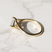 A gold ring with a asscher-cut clear diamond, resting on a white marble surface with soft shadows.