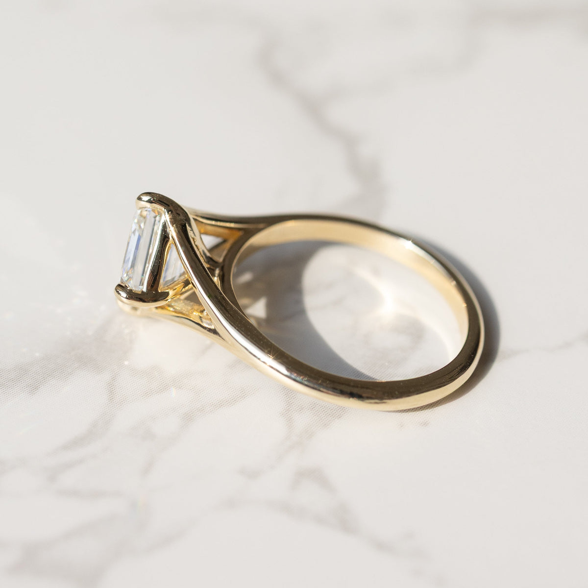 A gold ring with a asscher-cut clear diamond, resting on a white marble surface with soft shadows.