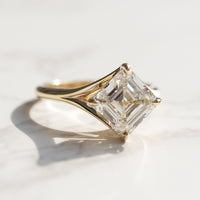 A yellow gold engagement ring with a large asscher diamond in the middle sitting on a white marble backdrop in the sun.