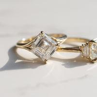 Two gold engagement rings resting on a light marble surface with soft shadows. The one in focus has a large asscher cut diamond kite set in a yellow gold setting.
