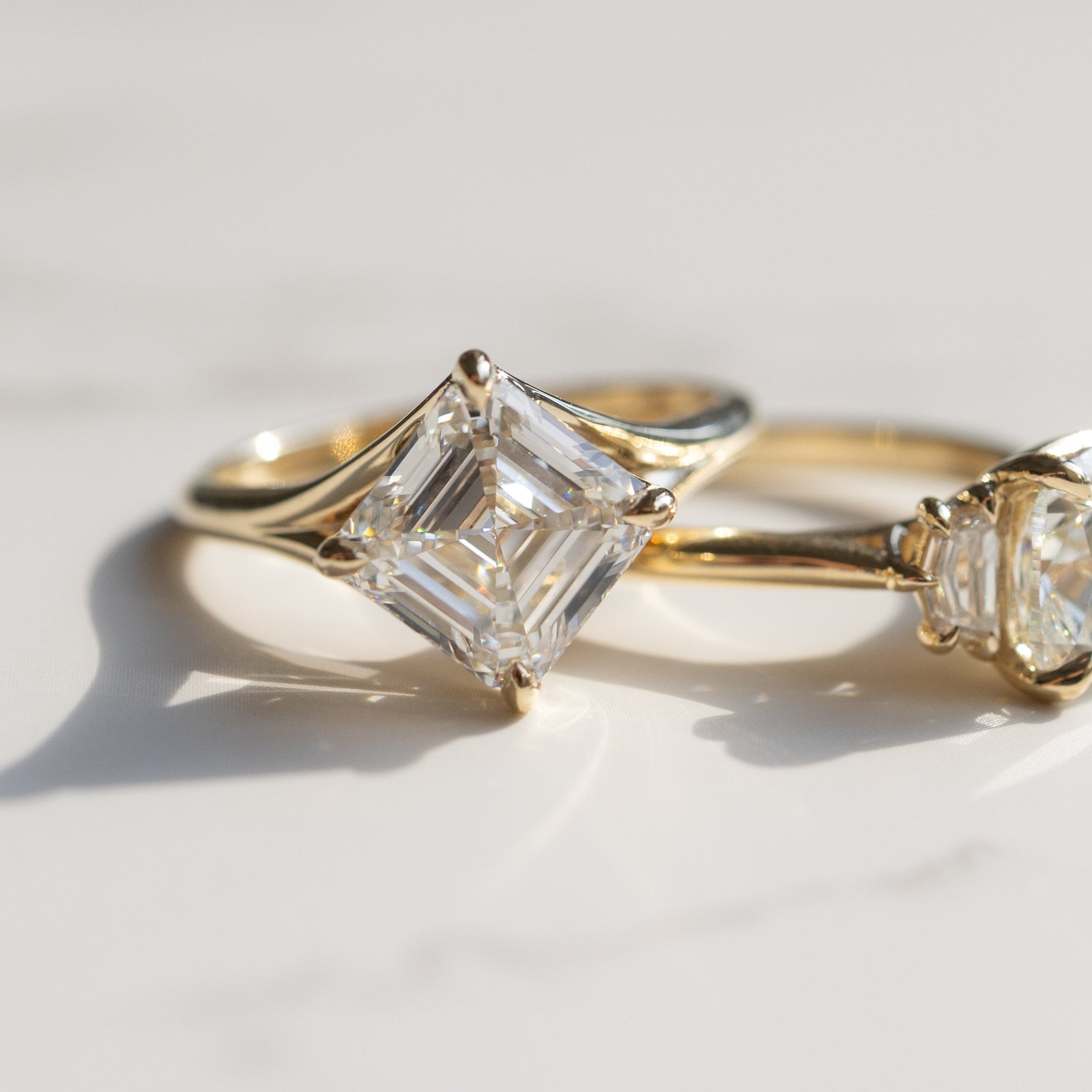 Two gold engagement rings resting on a light marble surface with soft shadows. The one in focus has a large asscher cut diamond kite set in a yellow gold setting.