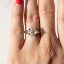 A close-up of a hand wearing a gold ring with an asscher cut diamond in the center. The hand has bright red nail polish, and the background is a clean, light marble surface.