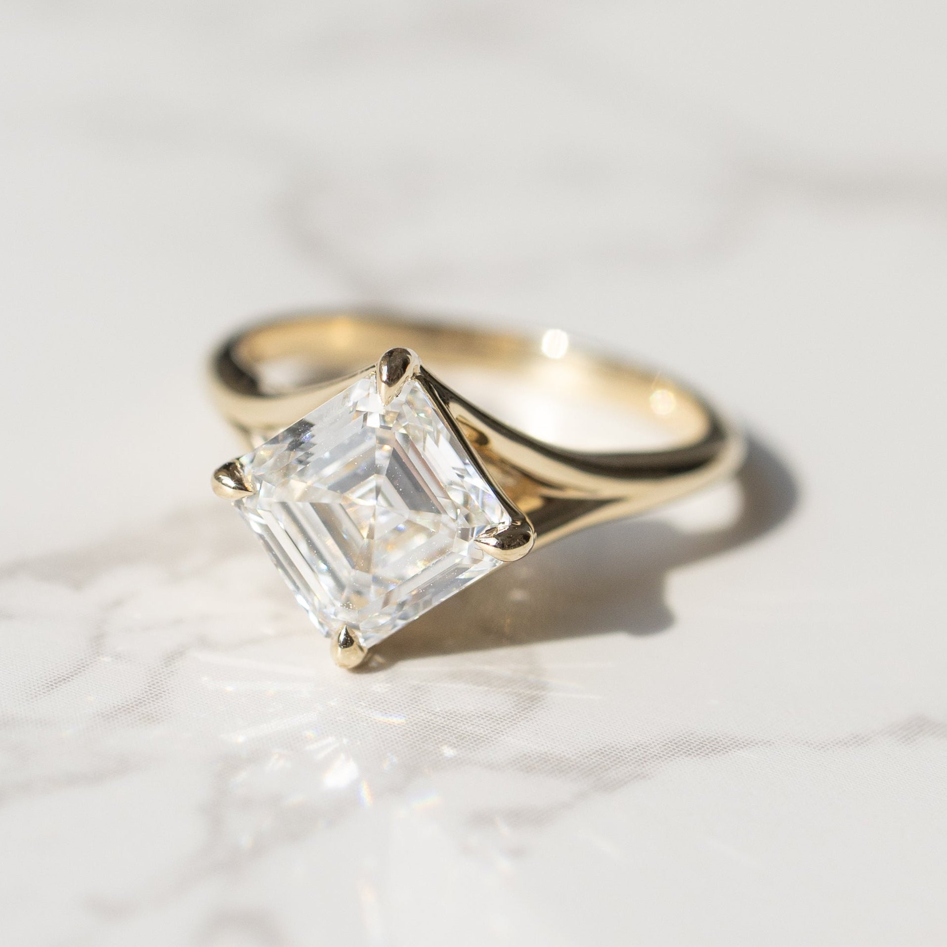 A yellow gold ring featuring a large, kite set asscher diamond in a split shank prong setting, resting on a white marble surface with soft shadows.