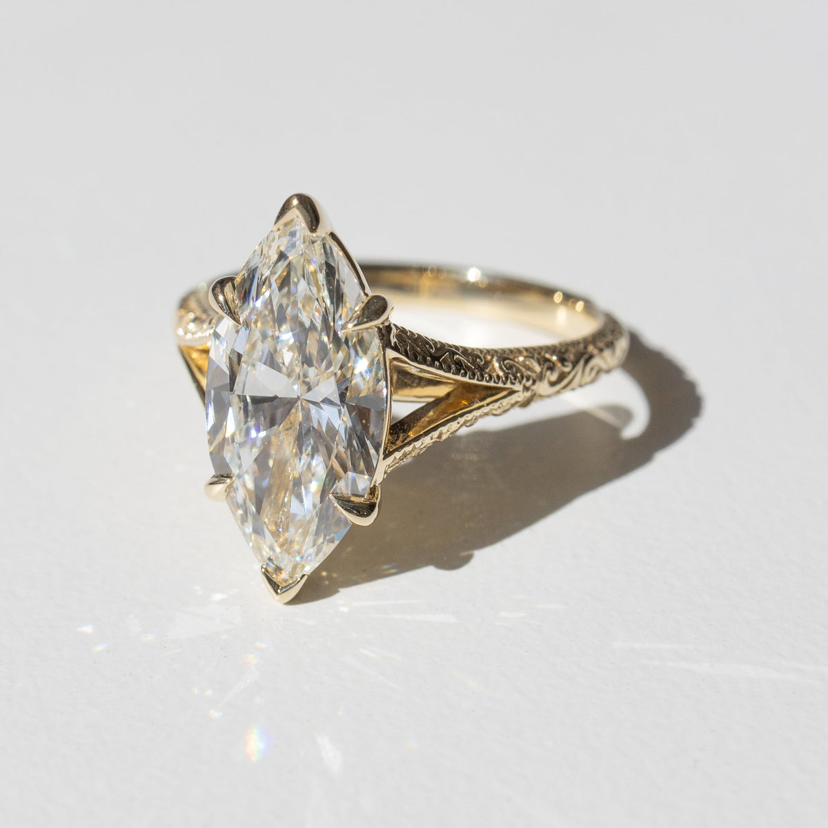 An intricate yellow gold engagement ring with a large marquise diamond in the center sitting on a white backdrop in the sun.