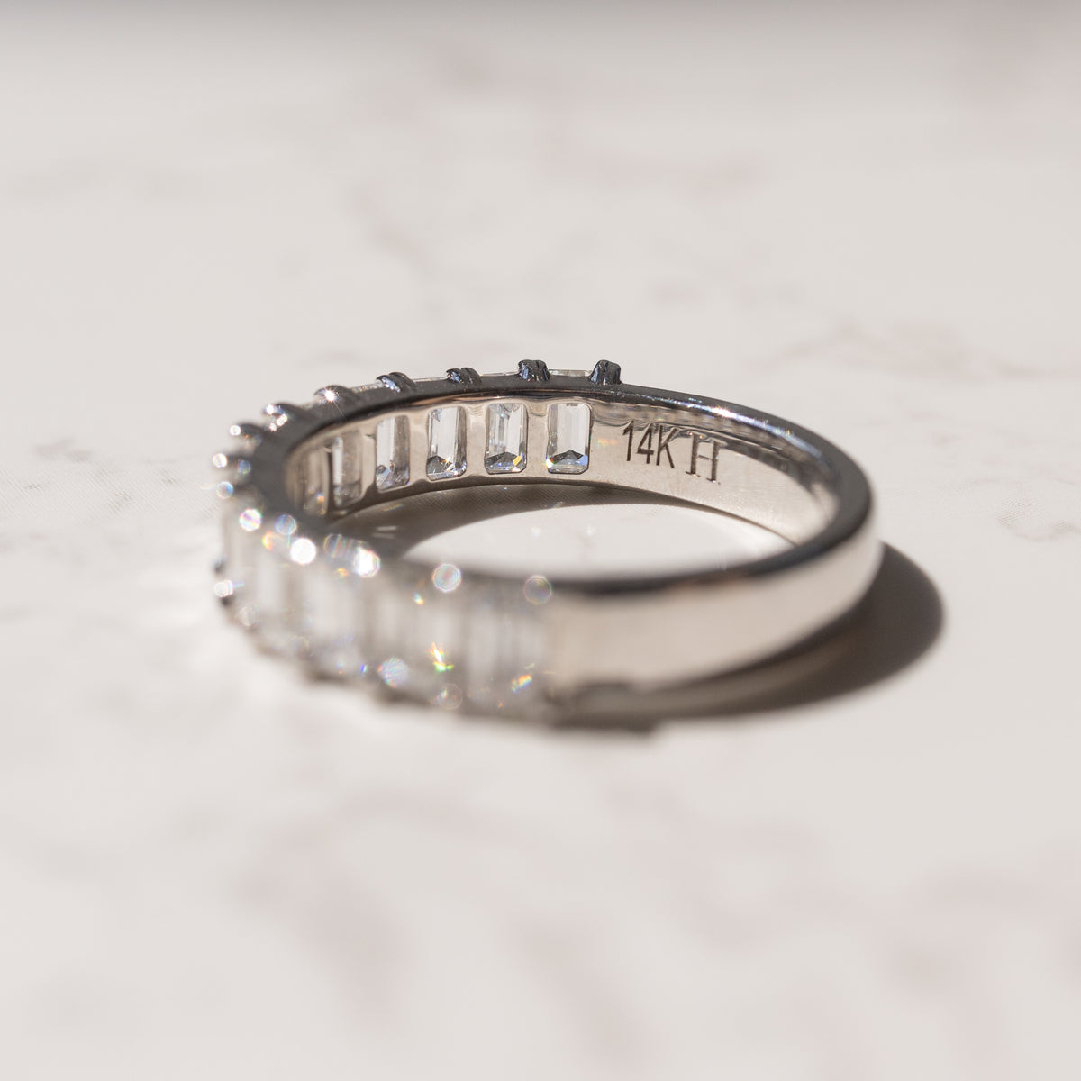 A white gold ring with emerald-cut clear diamonds, photographed on a light marble surface. The inside of the band is engraved with '14K'.