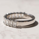 A diamond wedding band featuring a row of emerald-cut diamonds, photographed on a light marble surface with a soft shadow.