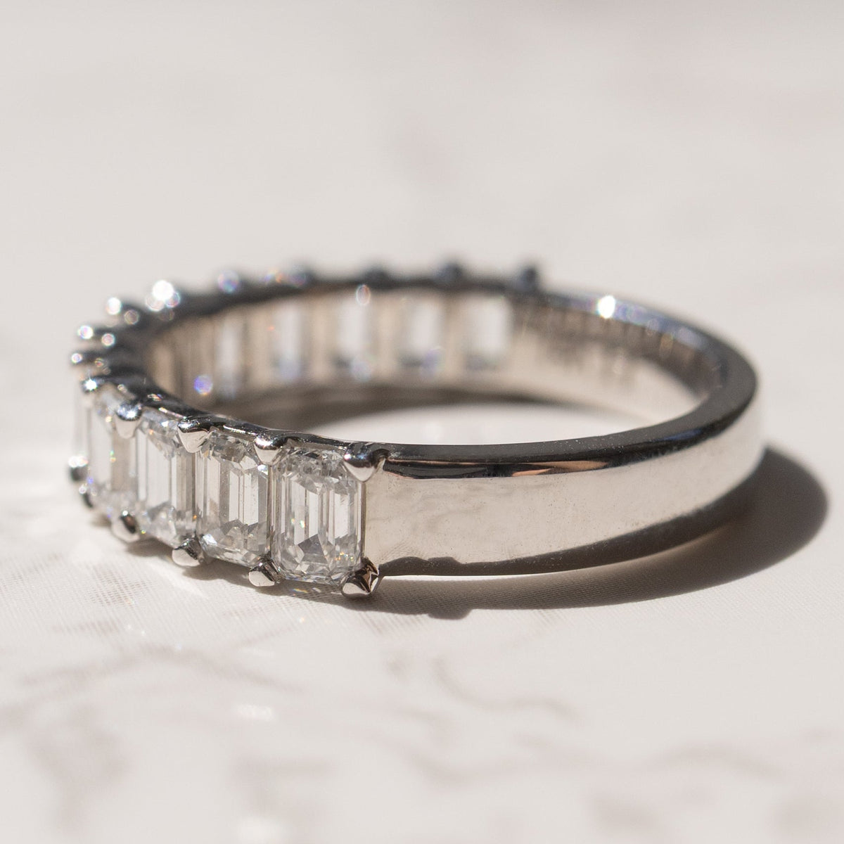 A diamond wedding band featuring a row of emerald-cut diamonds, photographed on a light marble surface with a soft shadow.