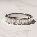 A white gold band ring featuring a row of emerald-cut diamonds, photographed on a light marble surface with a soft shadow.