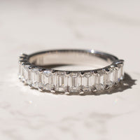 A white gold band ring featuring a row of emerald-cut diamonds, photographed on a light marble surface with a soft shadow.