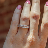 A close-up of a hand wearing a diamond wedding band with a row of emerald-cut diamonds. The hand has pink nail polish, and the background is a warm, blurred brown.