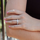 A close-up lifestyle shot of a person's hand resting on their elbow, wearing a chunky diamond ring with emerald-cut diamonds. The background is a soft-focus outdoor scene with green foliage and a dark railing.