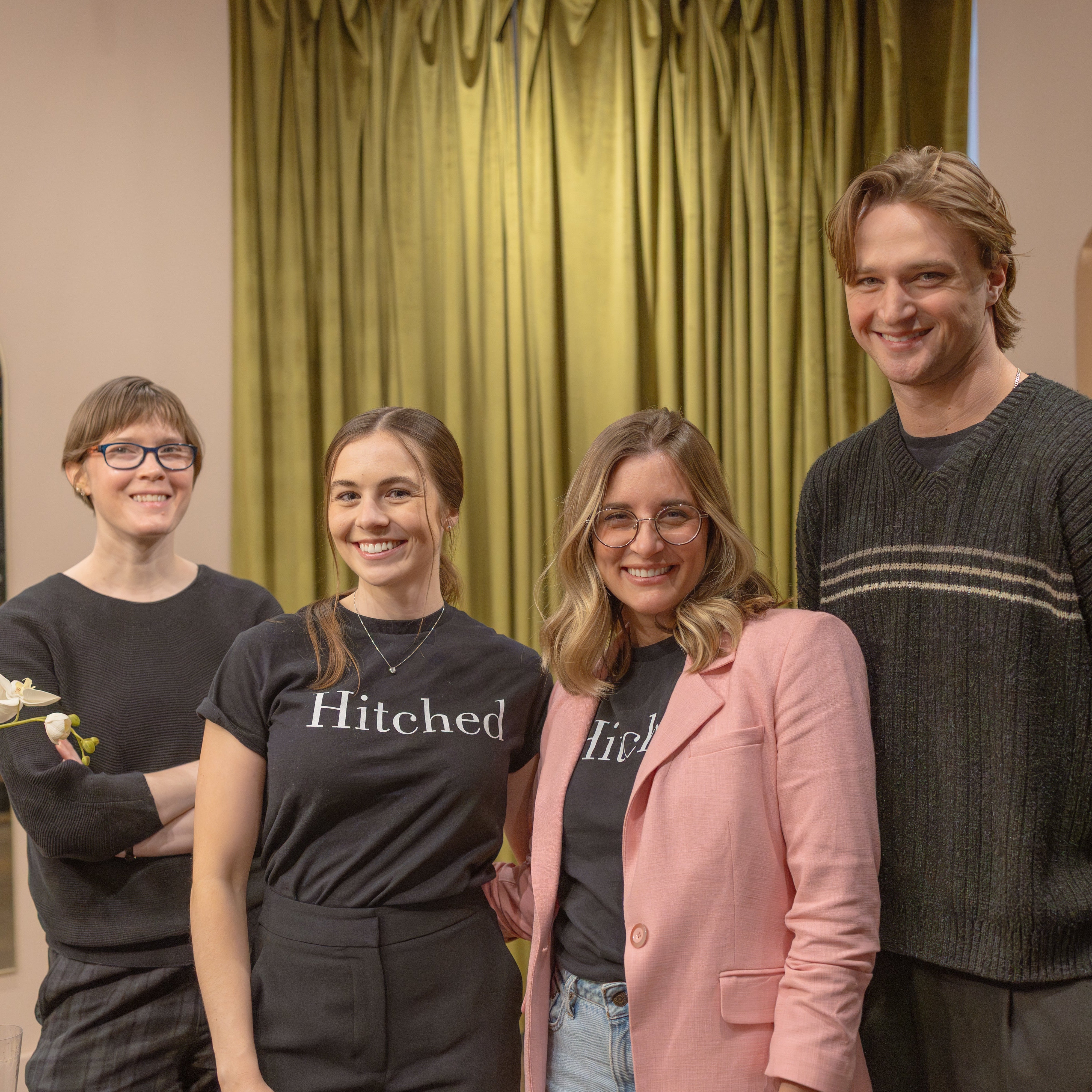 4 smiling people in a team group photo. They are wearing pink and black and standing in front of a green curtain.