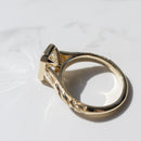 The cathedral profile of a yellow gold engagement ring with a bezel and engraving detail. The ring is sitting on a white marble backdrop.