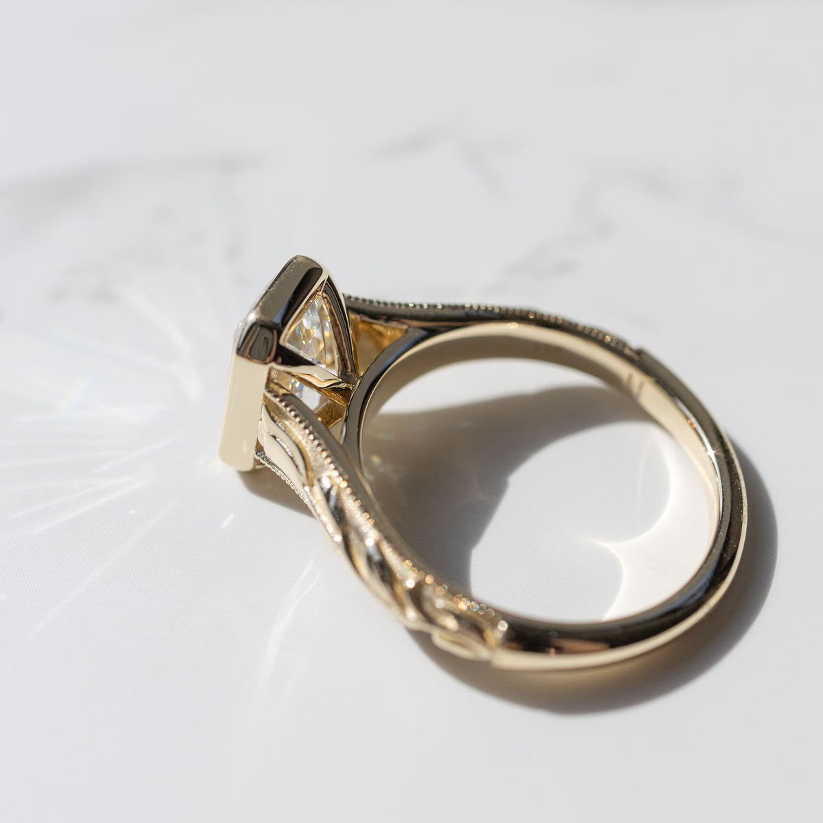 The cathedral profile of a yellow gold engagement ring with a bezel and engraving detail. The ring is sitting on a white marble backdrop.