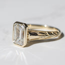 Macro shot of a emerald cut diamond engagement ring. In focus is a tulip detail along its yellow gold band.