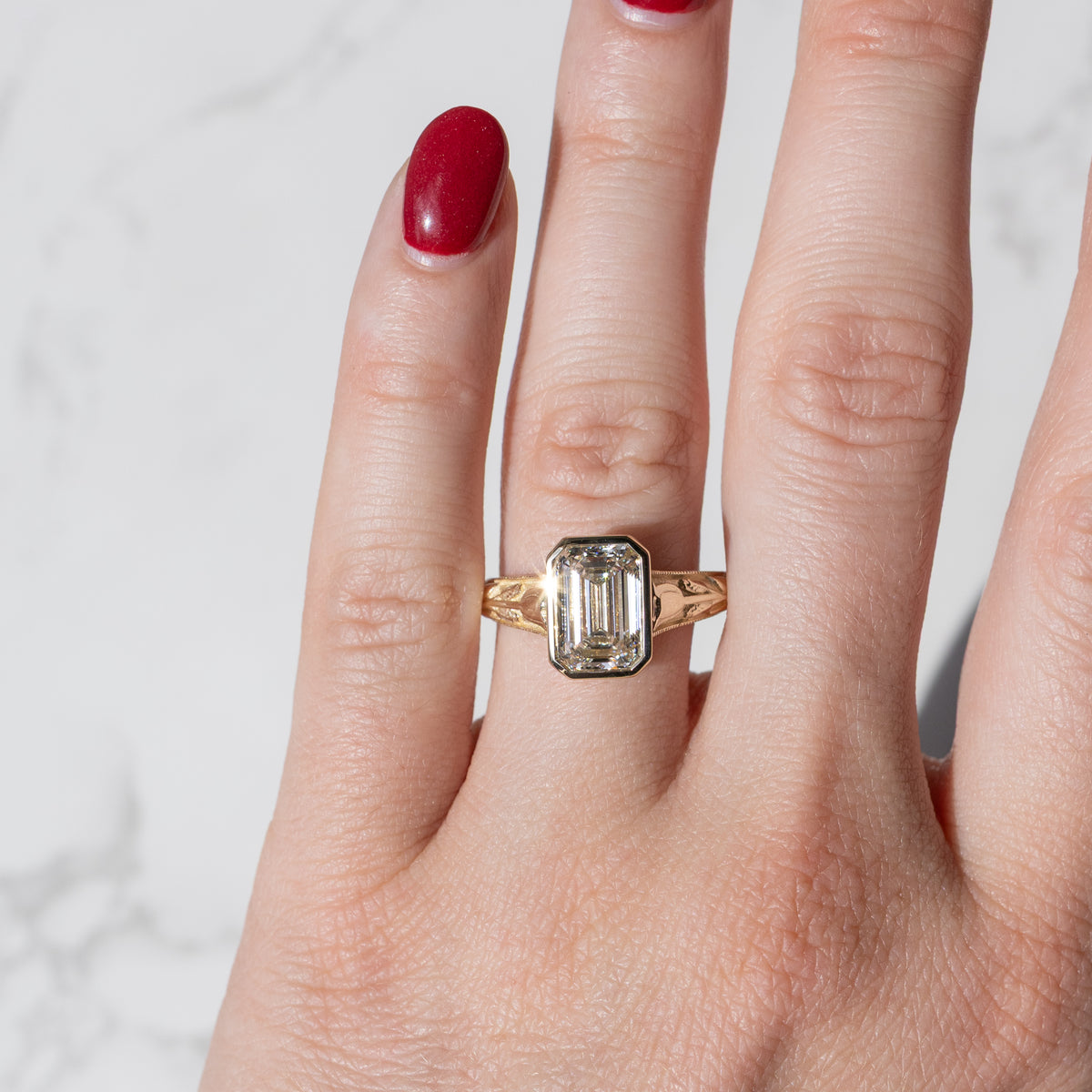 Product shot of an intricate yellow gold bezel engagement ring on a hand with bright red nail polish against a white marble backdrop.