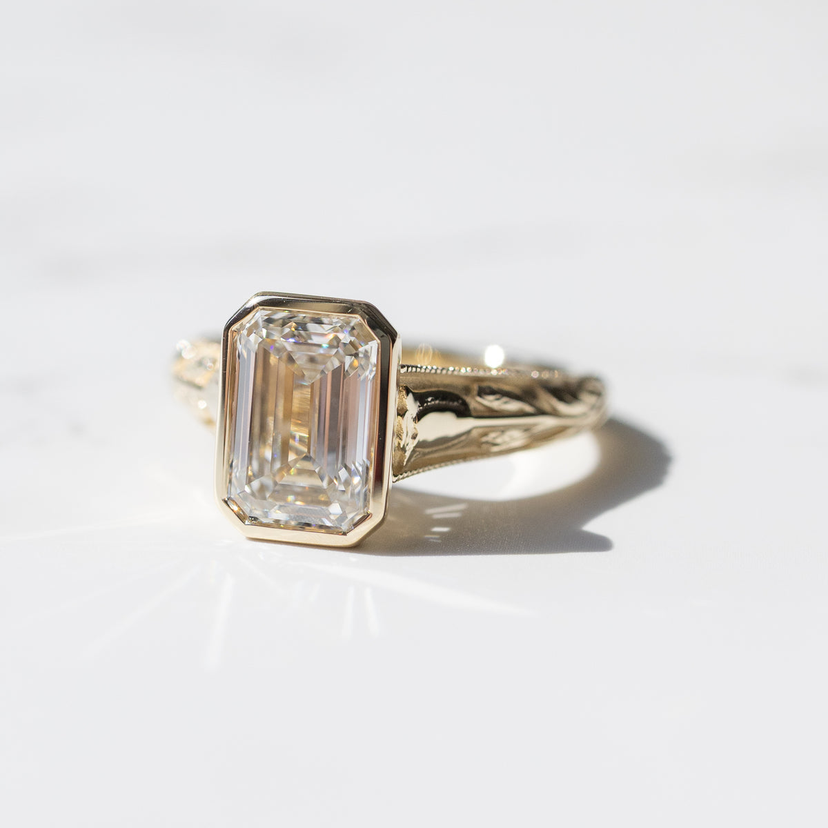 A large emerald cut diamond engagement ring sitting on a white marble background with soft shadows. The diamond is in a yellow gold bezel setting.