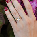 On-hand shot of a large emerald cut engagement ring on the left ring finger of a woman's hand. Behind the hand is blurry purple flowers and green leaves. The ring is yellow gold with intricate details on the band.