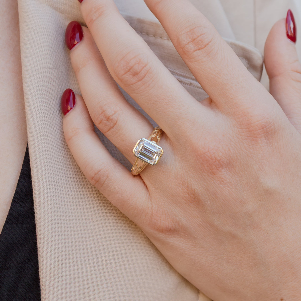 A woman's hand up against her trench coat. She has bright red nail polish and on her left ring finger is a large emerald cut diamond engagement ring with a bezel.