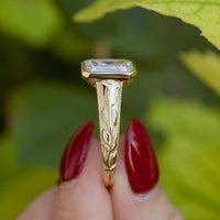 Close macro shot of the side profile of a chunky gold engagement ring. In focus is a tulip engraving going up the side of the band. Behind the ring is blurry green foliage.